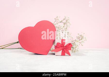 Red heart-shaped box and a small gift box tied with a red ribbon next to babys breath flowers on a light pink background. Valentines day concept. Stock Photo