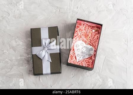 Gray gift box with silver ribbon and decorative heart ornament inside, surrounded by pink shredded paper on gray textured background. Valentines day c Stock Photo