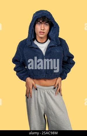 Fashionable young latin man posing in studio with blue furry cropped jacket and grey sweatpants on yellow background Stock Photo