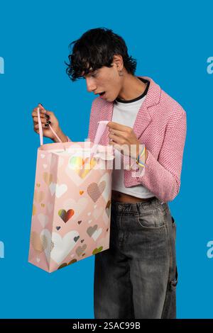 Young person opening a love-themed gift bag with a surprised expression, celebrating a special occasion on a vibrant blue background Stock Photo