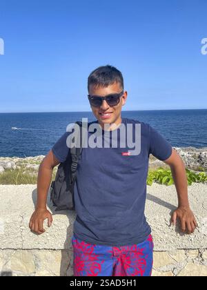 Young man standing against a wall and smiling at the camera, sea in the background, Italy Stock Photo