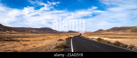 A long, winding road stretches through a vast, arid landscape with mountains in the background under a blue sky with scattered clouds. Stock Photo