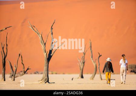 Back view of family mother and daughter walking among dead camelthorn trees surrounded by red dunes in Deadvlei in Namibia Stock Photo