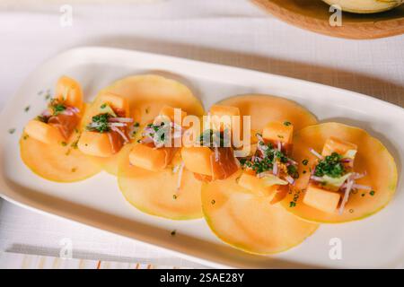 Sliced cantaloupe arranged on a white platter, topped with delicate slices of cured meat and fresh herbs, creating an elegant presentation at a dining Stock Photo