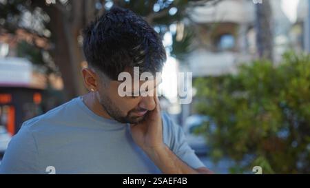Young hispanic man in a park setting touching his face, appearing thoughtful, with cityscape backdrop and natural ambiance, capturing an urban lifesty Stock Photo