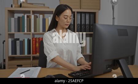 Young chinese woman typing on a computer in a modern office workspace with shelves full of books and documents in the background, portraying a profess Stock Photo