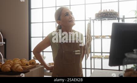 Woman smiling in a bakery, standing near pastries, wearing an apron, with shelves of baked goods in the background, in a brightly-lit shop with large Stock Photo