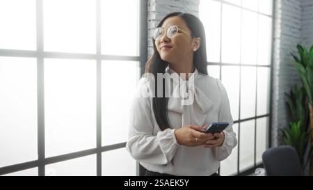 Woman smiling in bright office holding smartphone wearing glasses and white blouse standing near large window with plants adding a modern touch to the Stock Photo