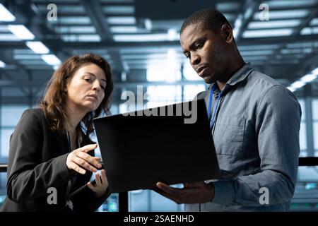 Team of engineers working in server farm, using laptops to analyze data. Teamworking employees examining infrastructure in data center, ensuring system integrity and security Stock Photo