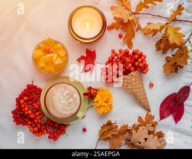 Autumn home cozy composition with cup of coffee, in a waffle cone, autumn leaves, burning candle and orange jam on a white knitted background. Stock Photo