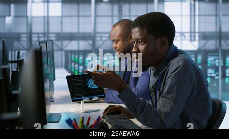 Cheerful coworkers in data center implementing AI predictive maintenance systems. Server room engineers using artificial intelligence to identify hardware failures impacting operations, camera A Stock Photo
