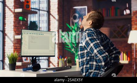 Software developer at computer coding, stretching joints after sitting at desk too long. IT expert in home office doing muscle exercises after feeling pain from incorrect posture, camera B Stock Photo