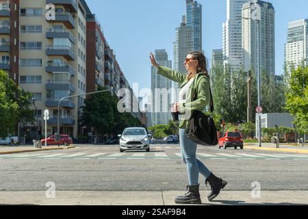 Young woman with a coffee cup and bag is hailing a cab on a busy city street, with modern buildings and traffic in the background during a sunny day Stock Photo