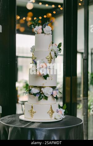 A beautifully crafted five-tier wedding cake stands elegantly on a round table, decorated with fresh flowers and gold accents, set in a modern venue f Stock Photo
