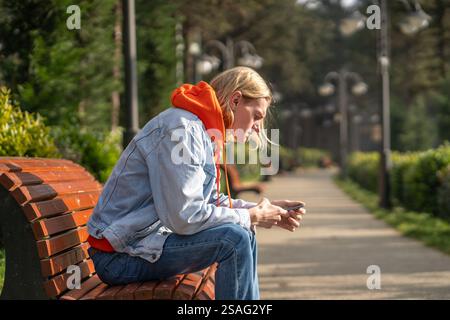 Thoughtful man sits on bench in public park, waiting for date with girl, writing message on phone Stock Photo