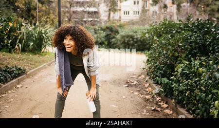 Woman in activewear taking a rest during a run. Aesthetic green park setting enhances the cheerful and calming atmosphere. Stock Photo