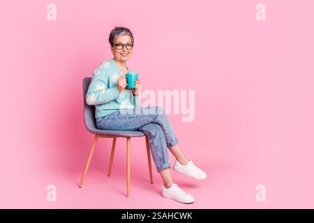 Stylish and mature woman in casual springwear with blue cardigan seated against a pink background holding a cup Stock Photo