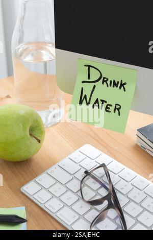 A simple yet effective image showing part of a computer screen on a desk, with a sticky note that says "Drink Water," serving as a reminder to stay hy Stock Photo