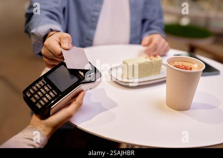 Close-up of hands making contactless payment in restaurant with credit card. Paper cup and dessert shown on table in background, creating cozy dining atmosphere Stock Photo