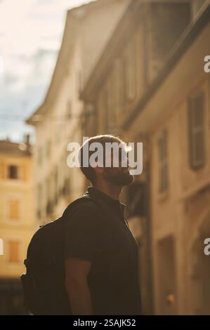 A young bearded man with a backpack and sunglasses walks through a historic European street, enjoying the golden sunlight and city atmosphere. Stock Photo