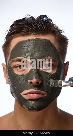 Man with smooth skin looking directly forward, and hand wearing black glove holding brush, mask covering most of face against white studio background. Stock Photo