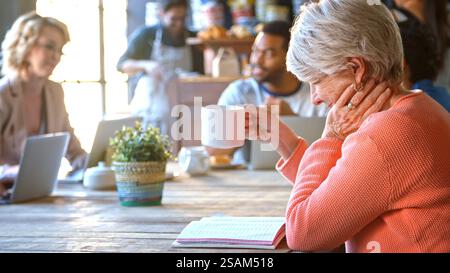 Senior Female Customer In Coffee Shop Sitting At Table With Drink Reading Notebook Stock Photo