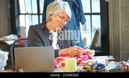 Senior Female Fashion Designer Using Laptop In Studio Checking Fabric Swatches Stock Photo