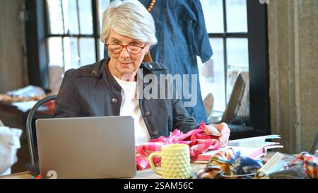 Senior Female Fashion Designer Using Laptop In Studio Checking Fabric Swatches Stock Photo