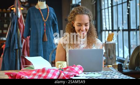 Young Female Fashion Designer In Studio Sitting At Desk Working On Laptop Stock Photo