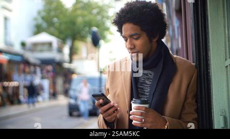 Young Man With Takeaway Coffee Checking Messages Or Social Media On City Street Using Mobile Phone Stock Photo