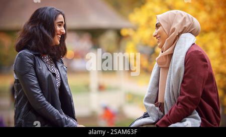 Two Young British Muslim Female Friends Sitting On Bench In Autumn Park Stock Photo