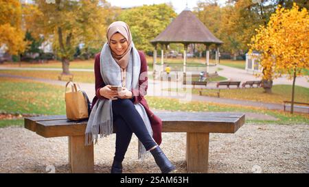 British Muslim Woman Sitting On Bench Using Mobile Phone To Check Messages And Social Media In Park Stock Photo