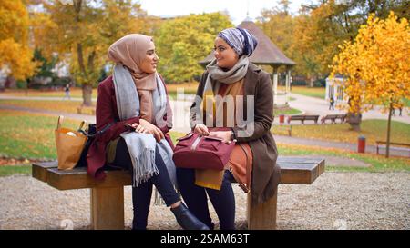 Two Young British Muslim Female Friends Sitting On Bench In Autumn Park Stock Photo