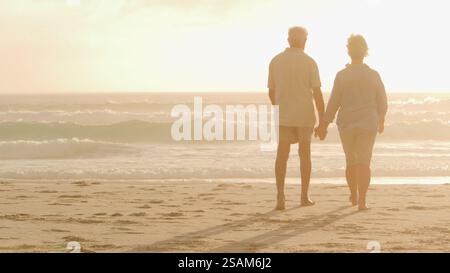 Rear View Of Senior Couple Holding Hands On Beach Vacation Watching Sun Set Over Ocean Stock Photo