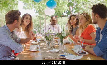 Group Of Multi-Cultural Young Friends Sitting Outdoors At Table Enjoying Cake And Hot Drink Together Stock Photo