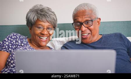 Senior Couple At Home Lying In Bed Having Video Call With Relatives On Laptop Stock Photo