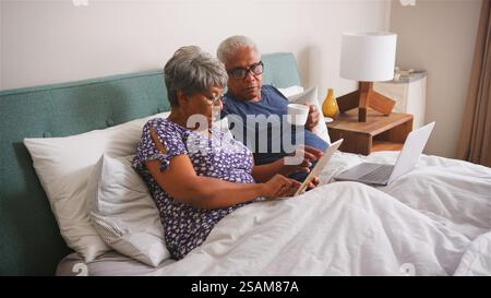 Senior Couple Lying In Bed At Home Using Digital Devices Stock Photo