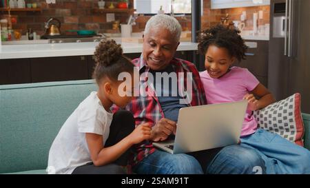 Grandfather Sitting On Sofa At Home With Granddaughters Using Laptop Together Stock Photo