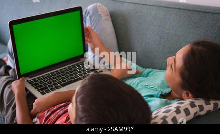 Rear View Shot Of Two Children Sitting On Sofa At Home Using Green Screen Laptop Computer Together Stock Photo