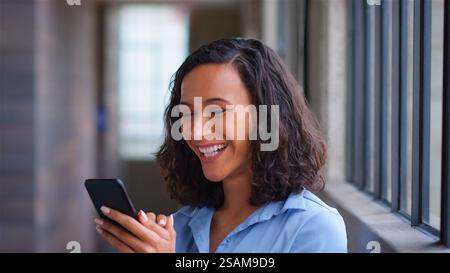 Smiling Businesswoman Standing By Office Window Using Mobile Phone Stock Photo
