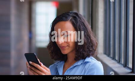 Smiling Businesswoman Standing By Office Window Using Mobile Phone Stock Photo