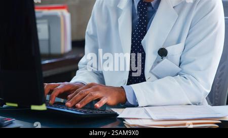 Close Up Of Male Doctor Wearing White Coat Behind Desk In Office Working At Computer Stock Photo
