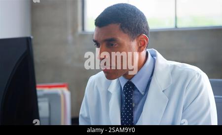 Male Doctor Wearing White Coat Sitting Behind Desk In Office Working On Computer Stock Photo