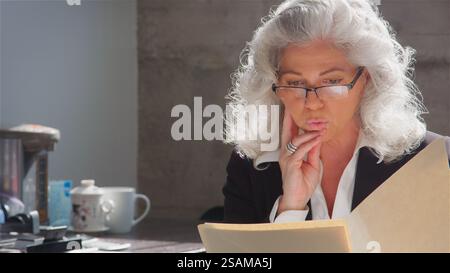 Mature Businesswoman Wearing Glasses Sitting At Desk In Office Reading Document Or File Stock Photo