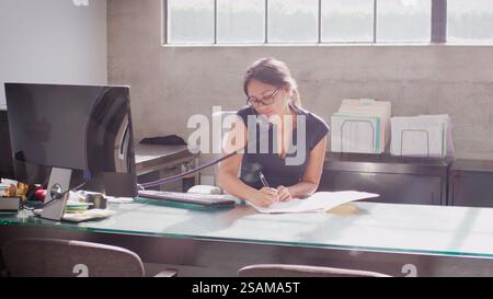 Businesswoman Wearing Glasses Sitting At Desk In Office Making Notes On Document Talking On Phone Stock Photo