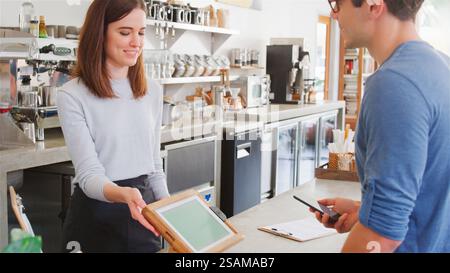 Female Worker Behind Counter Taking Contactless Payment From Male Customer With Phone In Coffee Shop Stock Photo