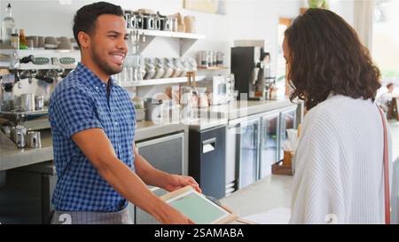 Male Worker Behind Counter Taking Contactless Payment From Female Customer In Coffee Shop Stock Photo