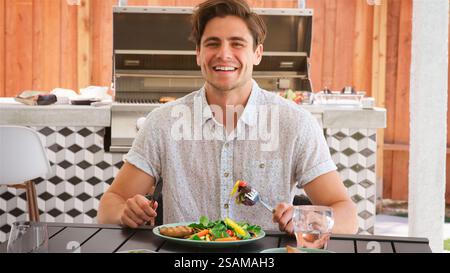 Portrait Of Young Man Outdoors At Home Sitting At Table Eating Barbecue Meal With Salad Stock Photo
