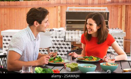 Young Couple Outdoors At Home Sitting At Table Eating Barbecue Meal With Salad Stock Photo