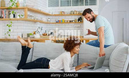 Young Couple At Home Woman On Sofa Using Laptop Computer With Man With Hot Drink In Kitchen Behind Stock Photo